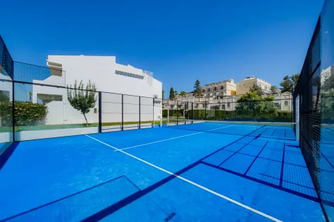 A blue padel court in the sun, white buildings behind it